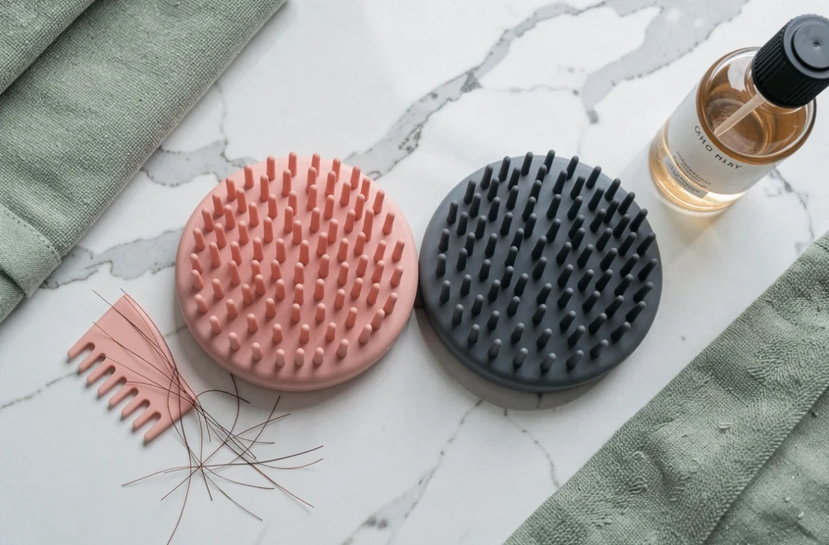Deux masseurs de cuir chevelu en silicone rose et gris posés sur un plan de travail en marbre blanc avec accessoires de soin capillaire